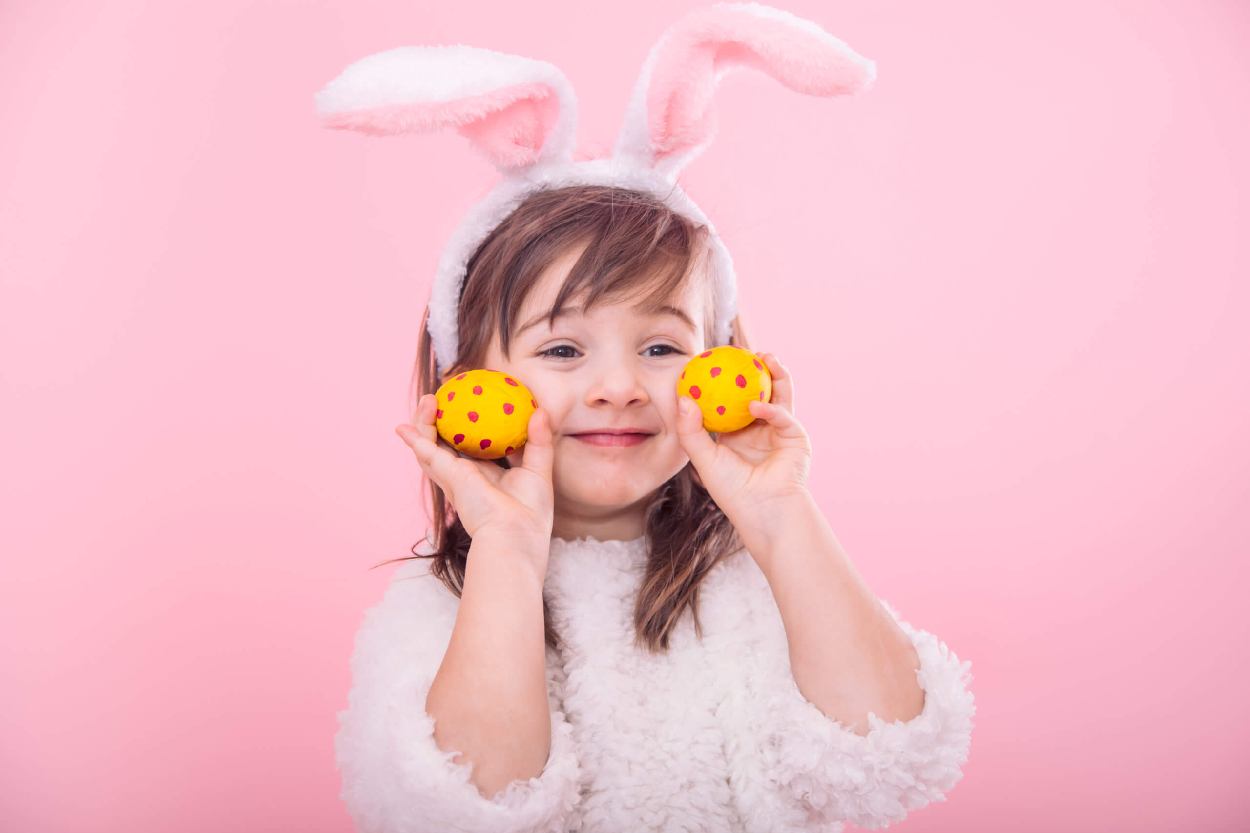 Portrait of a cute little girl with Bunny ears and yellow Easter eggs in red polka dots isolated on pink background