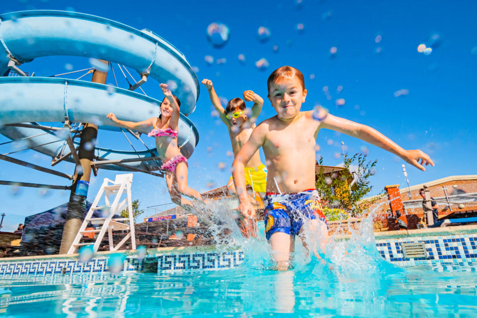 Three children jumping into an outdoor pool with a flume in the background